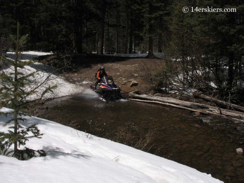 Frank Konsella snowmobiling across a stream. 